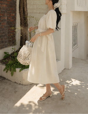 Woman in a white dress walking outdoors near a building with a bicycle.
