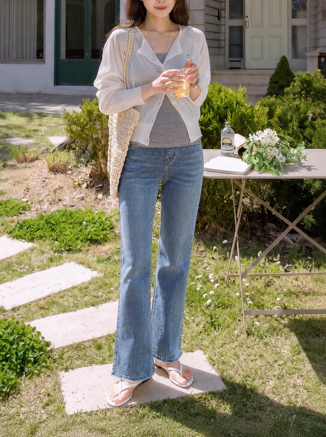 A woman stands outdoors in a grassy yard wearing light-wash blue flare jeans, a gray top, and a white button-up shirt, holding a drink. A table with flowers and a bottle is visible in the background.