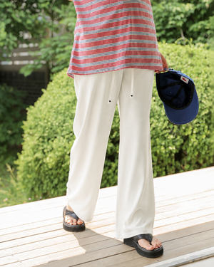A person standing outdoors on a wooden deck wearing off-white wide-leg drawstring pants, a pink and blue striped top, and black sandals, while holding a navy blue baseball cap.