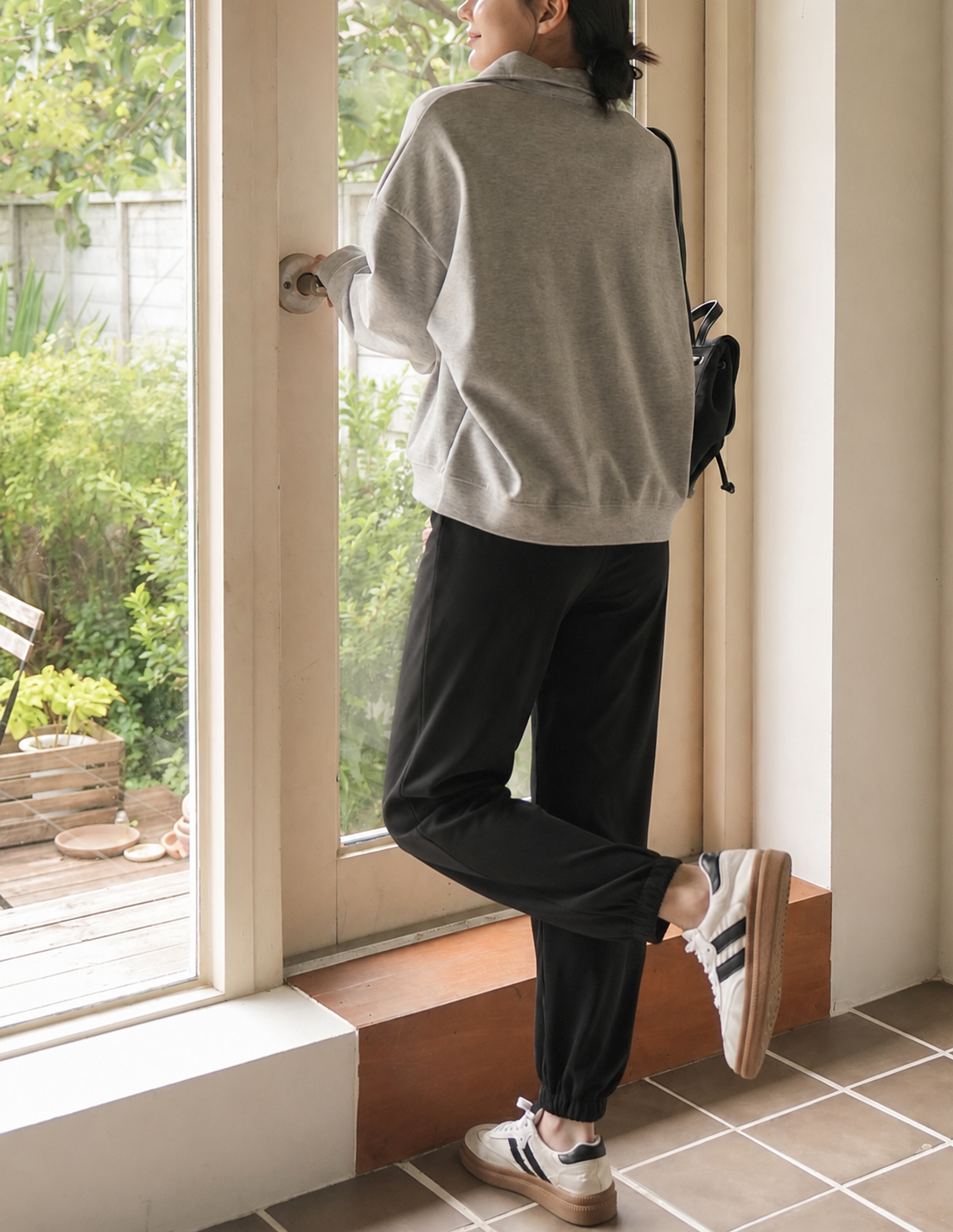 A person standing by a large window, wearing a light gray oversized sweatshirt, black jogger-style pants, and white sneakers with black stripes, carrying a small black shoulder bag.