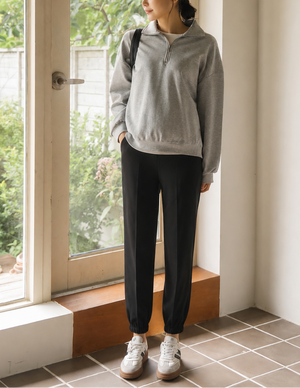 A woman stands indoors near a large window, wearing a light gray half-zip pullover, black jogger pants, and white sneakers.