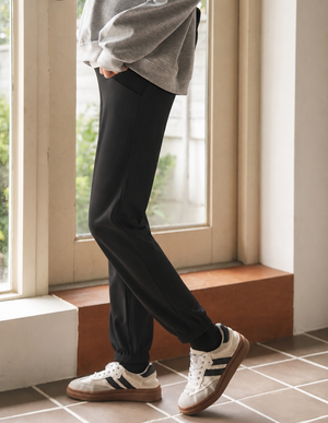 A person wearing black jogger-style pants with elastic cuffs, a loose gray sweatshirt, and white low-top sneakers with black stripes, standing indoors near a window.