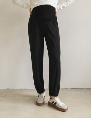 Black maternity pants with an elastic high-waist belly band and elastic ankle cuffs, shown on a model wearing white sneakers against a neutral studio background.