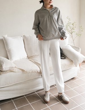 A person standing in a bright indoor room wearing a gray half-zip pullover and white jogger-style pants, with white sneakers, posed in front of a white sofa.