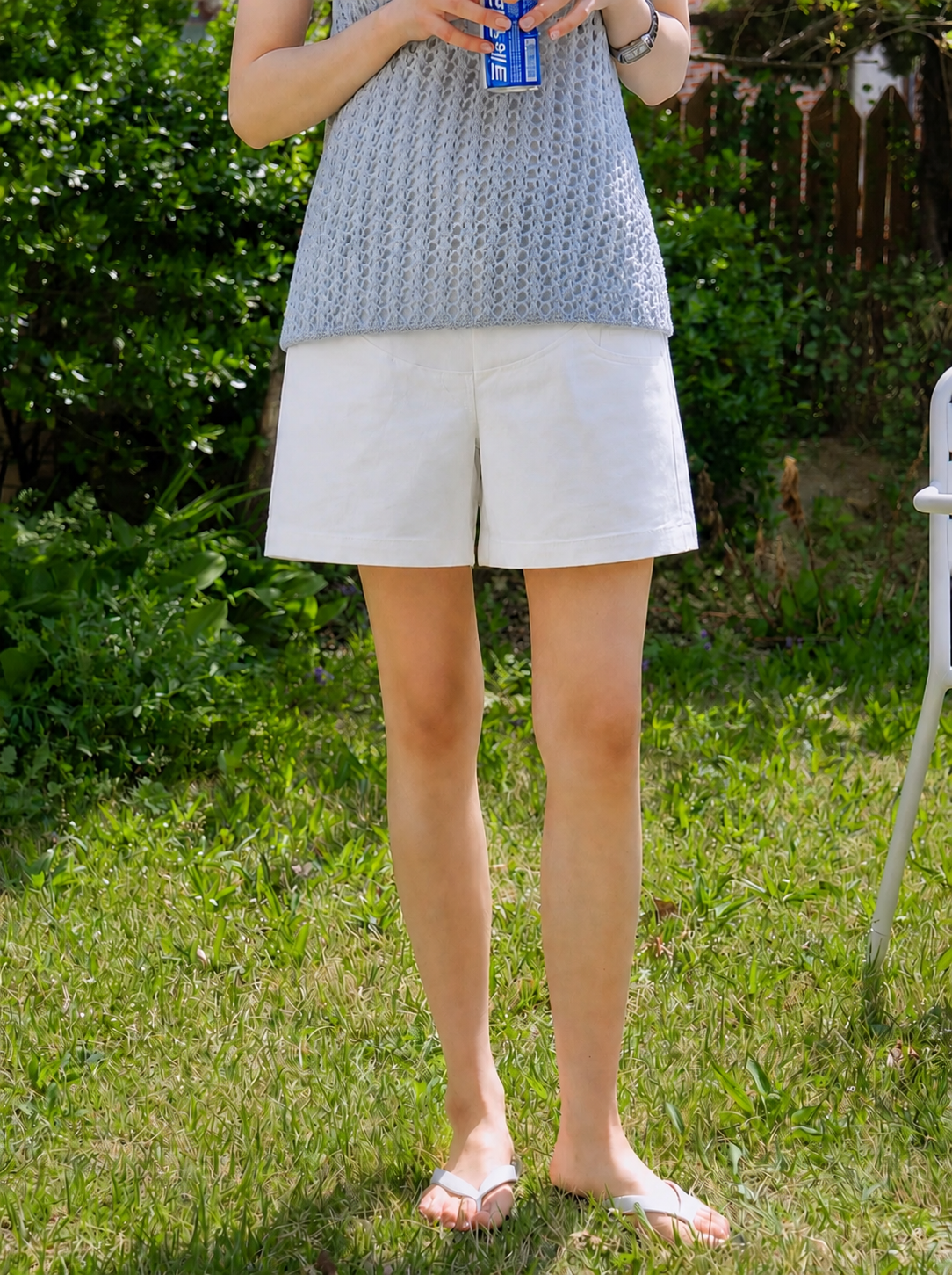 Person wearing a blue patterned top and white shorts standing on grass with greenery in the background