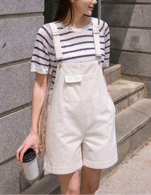 Woman wearing a striped shirt and white overalls standing against a gray stone wall.