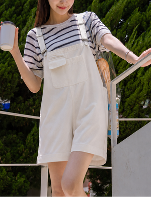 Woman in a striped shirt and white overalls holding a drink, standing outdoors with greenery in the background.