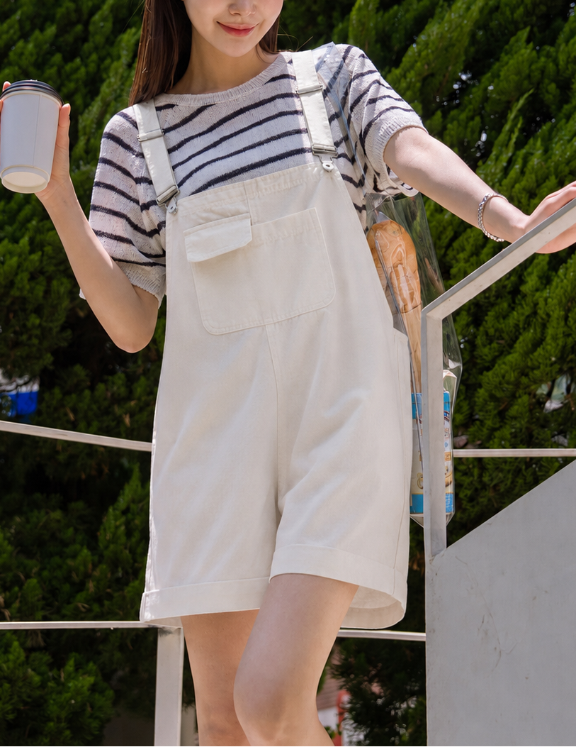 Woman in a striped shirt and white overalls holding a drink, standing outdoors with greenery in the background.