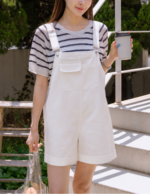 Woman in a striped shirt and white overalls holding a cup outdoors.