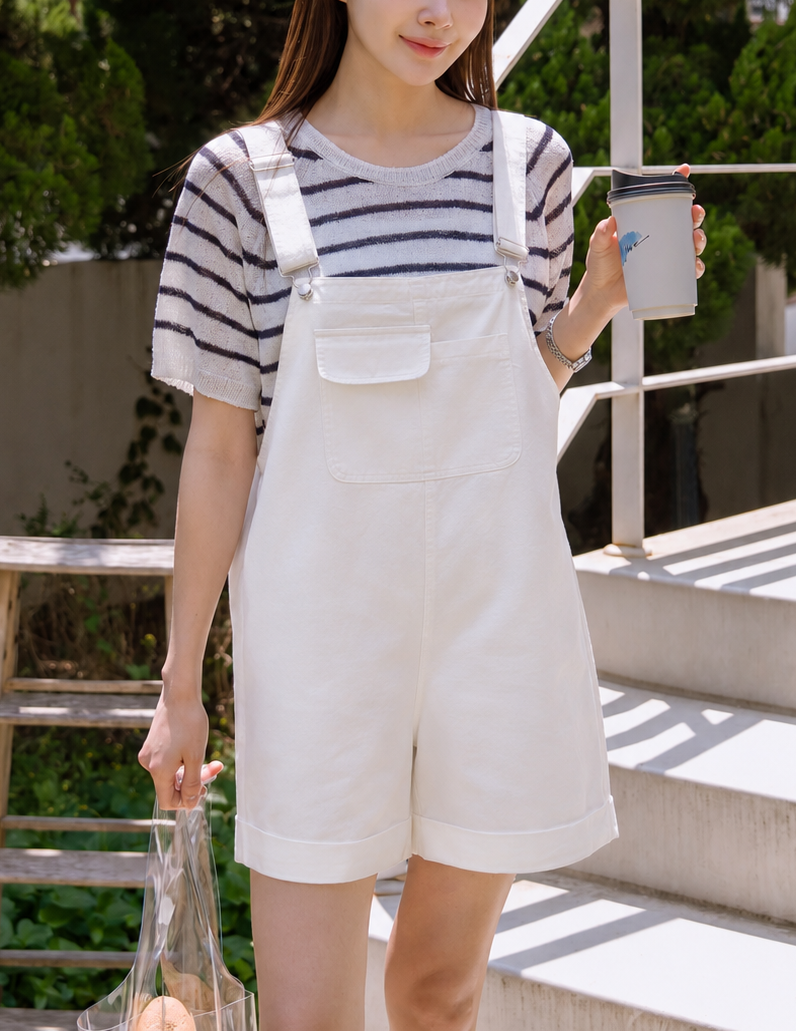 Woman in a striped shirt and white overalls holding a cup outdoors.