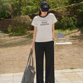 Person wearing a white t-shirt with text, black pants, and a cap, holding a striped bag outdoors.