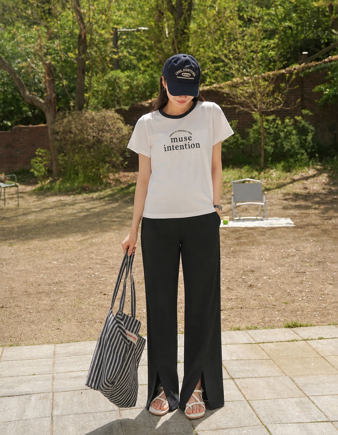 Person wearing a white t-shirt with text, black pants, and a cap, holding a striped bag outdoors.