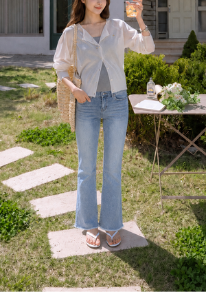 A woman stands outdoors in front of a house on a grassy lawn, wearing a sheer white long-sleeve top over a gray V-neck undershirt, light blue flared jeans, and flat sandals. She is holding a glass of iced coffee.