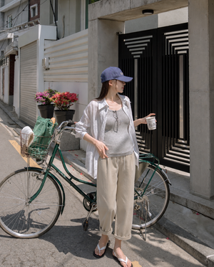 Woman standing next to a bicycle holding a drink, wearing a white shirt and beige pants, with a building and flowers in the background.