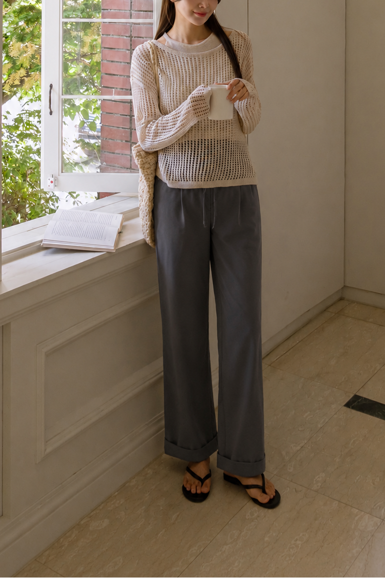 Woman standing by a window holding a phone in a room with light-colored walls and wooden floor.