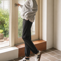 A woman stands in profile by a large window in a bright room, wearing a light gray oversized button-up shirt, dark charcoal pants, and white sneakers with black stripes.
