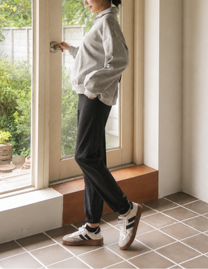 A woman stands in profile by a large window in a bright room, wearing a light gray oversized button-up shirt, dark charcoal pants, and white sneakers with black stripes.