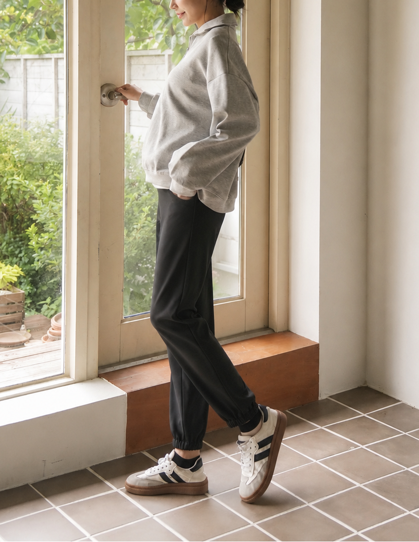 A woman stands in profile by a large window in a bright room, wearing a light gray oversized button-up shirt, dark charcoal pants, and white sneakers with black stripes.