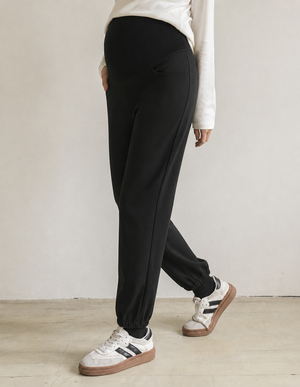 A person wearing black jogger-style pants with a white long-sleeve top and white sneakers with brown soles, standing against a plain light wall.