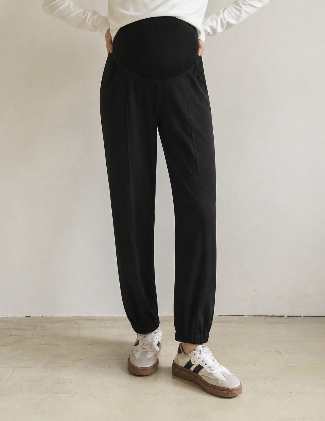 Black maternity pants with an elastic high-waist belly band and elastic ankle cuffs, shown on a model wearing white sneakers against a neutral studio background.