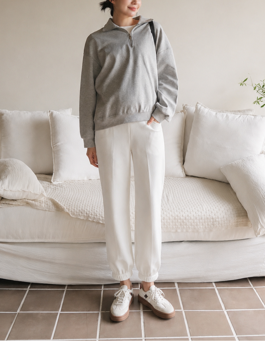 A woman stands in a softly lit room wearing a light gray half-zip pullover and off-white wide-leg sweatpants with white sneakers. She is posed in front of a cream-colored sofa with pillows and a tiled floor.